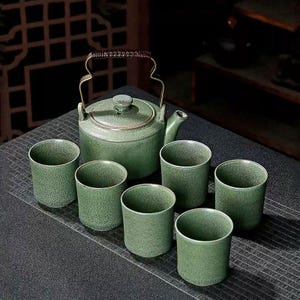 Puede incluir: Un juego de té de cerámica verde con una tetera con asa y tapa de metal, acompañado de seis tazas a juego. El juego tiene una textura moteada y está dispuesto sobre una superficie oscura.
