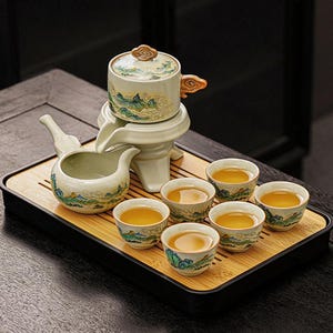 May include: A ceramic tea set featuring a teapot, server, and six small cups, decorated with a green and blue landscape design. The tea set is presented on a bamboo tray, with the cups filled with tea.