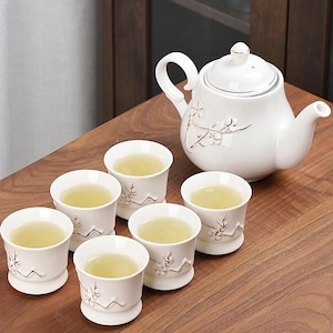 Puede incluir: Un juego de té de cerámica blanca con una tetera y seis tazas, todas llenas de té. La tetera y las tazas presentan un diseño floral con detalles dorados. El juego está dispuesto sobre una superficie de madera, creando una presentación serena y elegante.