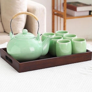 May include: A mint green ceramic tea set, including a teapot with a woven handle and six matching cups, presented on a dark wooden tray. The teapot has a rounded shape and a lid with a small knob. The cups are cylindrical.
