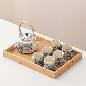 Może przedstawiać: Ceramiczny zestaw do herbaty z czajniczkiem i sześcioma filiżankami, z niebieskimi wzorami kwiatowymi na białym tle. Czajniczek ma bambusowy uchwyt i stoi na drewnianej tacy. Filiżanki są ułożone wokół czajniczka.