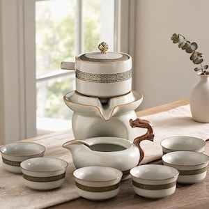 May include: A ceramic tea set featuring a white teapot with a gold Greek key pattern and a gold finial, a matching tea pitcher with a brown dragon-shaped handle, and six small white tea cups decorated with a matching gold Greek key band.