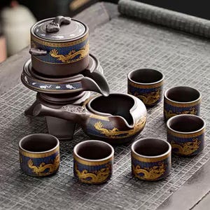 Może przedstawiać: Brązowy ceramiczny zestaw do herbaty ze złotym wzorem smoka na niebieskim tle. Zestaw zawiera czajniczek, naczynie do serwowania, podgrzewacz do herbaty i sześć małych filiżanek. Zestaw stoi na szarej powierzchni.