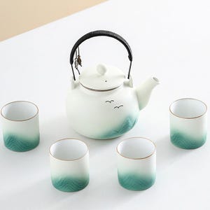 May include: A white ceramic teapot with a black handle and four matching cups. The teapot and cups feature a teal and white gradient design with a mountain and bird motif. The cups have a gold rim.