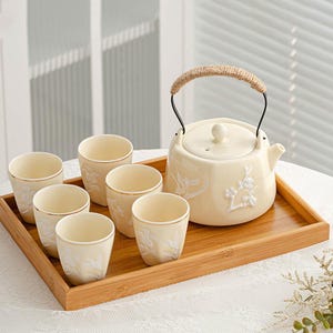 Puede incluir: Un juego de té color crema con seis tazas, una tetera y una bandeja de madera. La tetera tiene un asa tejida y detalles florales en relieve. Las tazas tienen bordes dorados y diseños florales. El juego está dispuesto sobre una bandeja de bambú.