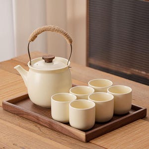 Pode incluir: Bule de chá de cerâmica creme com alça de corda e tampa de madeira, acompanhado por seis xícaras de chá combinando. O conjunto é apresentado em uma bandeja de madeira escura, perfeito para servir chá. Design minimalista.