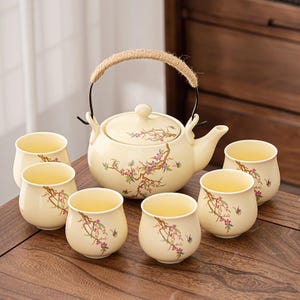 Puede incluir: Juego de té color crema con una tetera con asa de cuerda y seis tazas a juego. La tetera y las tazas están adornadas con un diseño floral en rosa y marrón. El juego se exhibe sobre una superficie de madera.
