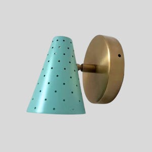 May include: A vintage-style wall sconce with a teal cone-shaped shade perforated with small holes. The sconce features a brass-colored circular backplate and a connecting arm. The design is reminiscent of mid-century modern lighting.