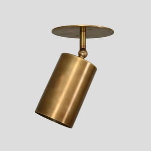 May include: A brass-coloured, cylindrical ceiling spotlight with a circular mounting plate. The spotlight is adjustable with a ball joint. The fixture has a warm, metallic finish, suitable for interior lighting. The spotlight is designed to direct light.
