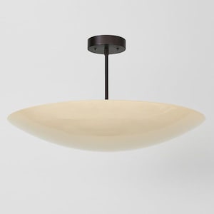 Puede incluir: Una moderna lámpara de techo de color crema con un vástago y dosel marrón oscuro. La lámpara tiene una pantalla ancha, poco profunda y de forma ovalada, creando un diseño minimalista y elegante. La lámpara está suspendida del techo.