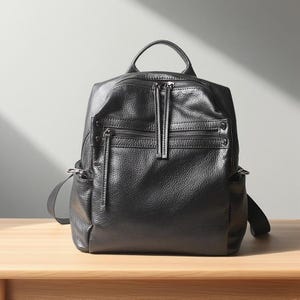 Può includere: Zaino in pelle nera con più scomparti con cerniera e una maniglia superiore. La borsa ha una superficie strutturata e spallacci regolabili. Lo zaino è appoggiato su una superficie di legno.