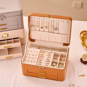 May include: A tan leather jewelry box with multiple compartments and drawers, open to display necklaces, rings, and earrings. Two other jewelry boxes are stacked behind it. A gold-framed mirror and perfume bottle are in the background.