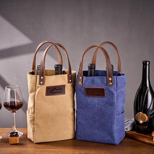 Puede incluir: Dos bolsas de lona para botellas de vino, una beige y otra azul, con asas de cuero marrón. Cada bolsa tiene un parche de cuero marrón y contiene una botella de vino. También se ven dos copas de vino y una botella de vino.