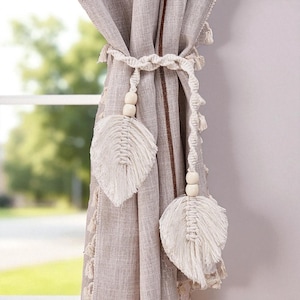 May include: Beige macrame curtain tiebacks with leaf-shaped accents and wooden beads. The tiebacks are wrapped around a beige curtain with a brown stripe. The curtain is hanging in front of a window.