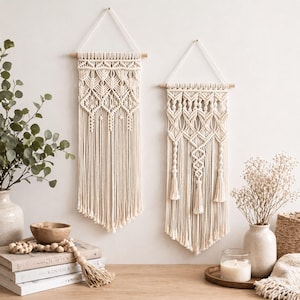 Könnte beinhalten: Zwei cremefarbene Makramee-Wandbehänge mit geometrischen Mustern und Fransen. Jedes Stück ist an einem Holzdübel und einer weißen Kordel aufgehängt. Die Makramee-Kunst wird vor einer weißen Wand ausgestellt und schafft eine Boho-Ästhetik.