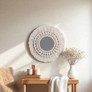 May include: A round, cream-colored macrame mirror with a fringe border. The mirror has a central reflective surface surrounded by a pattern of wooden beads. The mirror is hanging on a light-colored wall. A wooden table below holds candles and a vase.