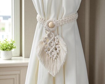 May include: Cream-colored macrame curtain tieback with a leaf design. The tieback features a woven rope band, a wooden button, and a decorative leaf shape with intricate knotting and fringe. The tieback is holding a white curtain.