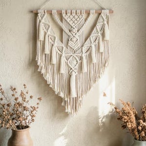 May include: Cream-coloured macrame wall hanging with a geometric design. The textile art piece features intricate knotting, tassels, and a wooden dowel for hanging. This bohemian-style decor adds texture and warmth to any space.