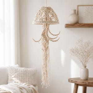 May include: A cream-colored macrame pendant light fixture. The lamp features a rounded shade with intricate woven patterns and long, dangling cords. The design evokes a jellyfish shape, adding a bohemian touch to interior decor. The light fixture is suspended from the ceiling.