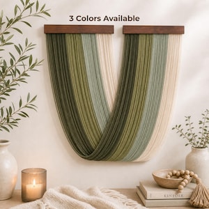 Op de afbeelding: Wanddecoratie met een houten bovenkant en groene en crèmekleurige garenstrengen. De tekst "3 Colors Available" is zichtbaar. De afbeelding bevat vazen, een kaars en boeken, wat een gezellige sfeer creëert.