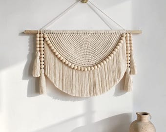 Puede incluir: Colgante de pared de macramé color crema con una clavija de madera y detalles de cuentas. El diseño presenta un patrón semicircular con largas franjas y borlas a cada lado. Una pieza decorativa para la decoración del hogar.