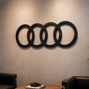 May include: A matte black wall-mounted sign featuring four interlocking circular rings arranged in a horizontal row. The design consists of four identical circles linked together, creating a continuous chain-like structure mounted on a wall.