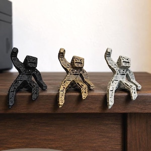 Op de afbeelding: Drie kleine, gearticuleerde figuurtjes met een geometrische, rasterachtige structuur. Eén is matzwart, één is metallic goud en één is metallic zilver. Elk figuurtje heeft een vierkant hoofd en ledematen in een zittende houding op een rand.
