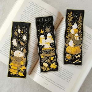 May include: Three black bookmarks with gold accents and illustrations. One shows ghosts with books and leaves, another has ghosts in a teacup, and the third features ghosts on pumpkins. The bookmarks are rectangular and rest on an open book.