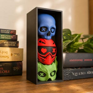 May include: A vertical black rectangular display box containing three stacked decorative objects: a blue skull with an X and heart eye, a red helmet with a heart and X eye, and a light green mask with an X and heart eye, all arranged in a column.
