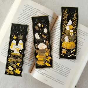 May include: Three black bookmarks with gold illustrations of ghosts, pumpkins, and autumn leaves. One bookmark features ghosts in a teacup, another has a cat on books, and the third shows ghosts on pumpkins. The bookmarks are on an open book.