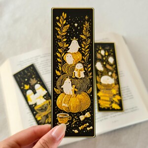 May include: Black and gold bookmark featuring a stack of pumpkins with ghost characters. The bookmark has a gold border and includes a teacup and candies. The design is set against a black background with gold accents.