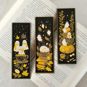 May include: Three black bookmarks with gold accents and whimsical illustrations. One features ghosts in scarves over a teacup, another a cat on books, and the last ghosts on pumpkins. The bookmarks are rectangular.