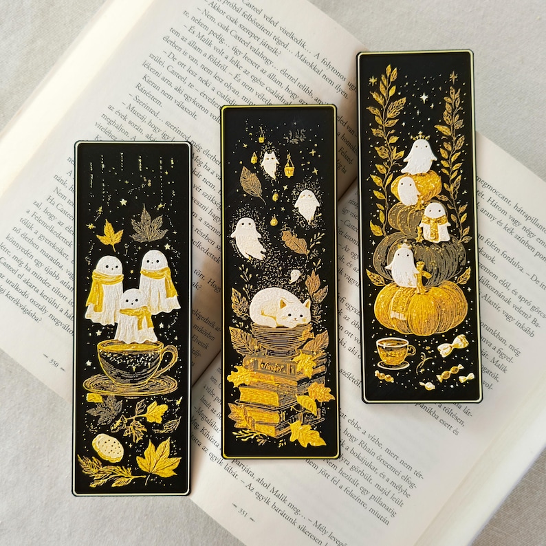 May include: Three black bookmarks with gold accents and whimsical illustrations. One features ghosts in scarves over a teacup, another a cat on books, and the last ghosts on pumpkins. The bookmarks are rectangular.