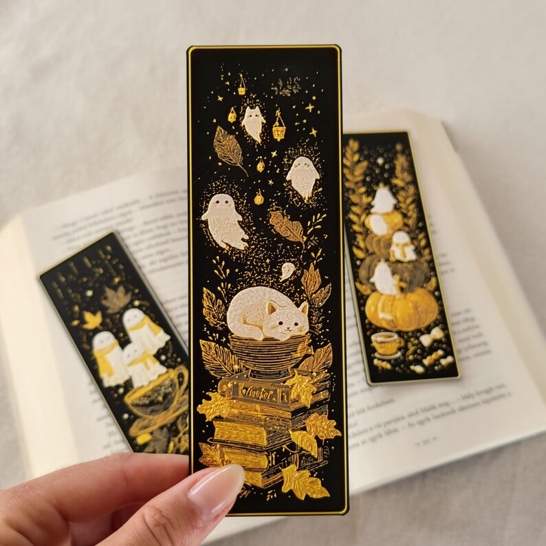 May include: Black rectangular bookmarks with gold illustrations. One bookmark features a cat on books, ghosts, and leaves. Another shows pumpkins and ghosts. The third bookmark has ghosts in a teacup. All are on a white surface.