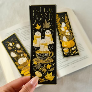May include: Three black and gold bookmarks with autumn-themed illustrations. One bookmark features three ghosts in scarves above a teacup. Another shows a stack of pumpkins and ghosts. The third has a sleeping animal.