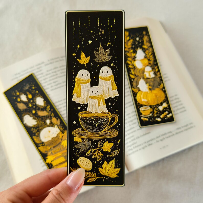 May include: Three black and gold bookmarks with autumn-themed illustrations. One bookmark features three ghosts in scarves above a teacup. Another shows a stack of pumpkins and ghosts. The third has a sleeping animal.