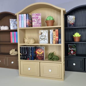 Peut inclure: Une petite étagère beige avec un dessus en arc, remplie de minuscules livres, une plante succulente en pot et des figurines décoratives. Les livres ont des titres comme "It Ends With Us" et "Cruel Prince". Deux tiroirs sont en bas. Des étagères marron et noires similaires sont en arrière-plan.