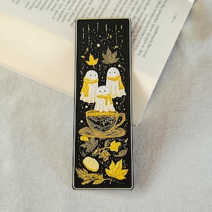 May include: A black bookmark featuring three ghost figures with yellow scarves, floating above a teacup. The design includes gold-colored leaves and stars, with a white border. The bookmark is resting on an open book.