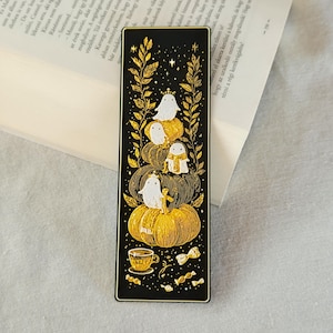 May include: Black bookmark with a gold design featuring ghosts on pumpkins, a teacup, and candies. The bookmark has a rectangular shape with rounded corners. The design is set against a black background with gold accents.