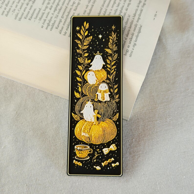 May include: Black bookmark with a gold design featuring ghosts on pumpkins, a teacup, and candies. The bookmark has a rectangular shape with rounded corners. The design is set against a black background with gold accents.