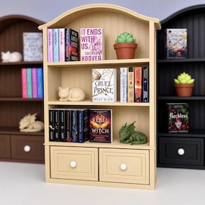 Può includere: Una libreria in miniatura in legno color beige chiaro con tre ripiani e due cassetti inferiori. I ripiani sono pieni di libri in miniatura, inclusi titoli come It Ends With Us, The Cruel Prince e Rebel Witch, insieme a una piccola pianta grassa in vaso, una statuetta di un gatto che dorme e un drago verde.