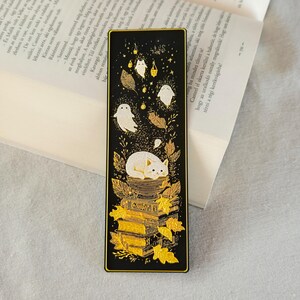 May include: Black bookmark with gold accents featuring a white cat, ghosts, leaves, and books. The bookmark has a rectangular shape with a gold border. The design includes a stack of books and falling leaves.