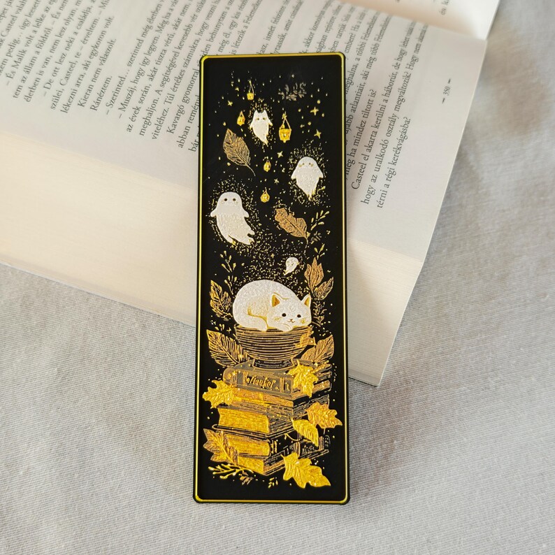 May include: Black bookmark with gold accents featuring a white cat, ghosts, leaves, and books. The bookmark has a rectangular shape with a gold border. The design includes a stack of books and falling leaves.