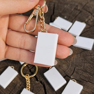 Op de afbeelding: Een kleine, rechthoekige, effen witte bedel in de vorm van een boek, bevestigd aan een goudkleurige metalen sleutelhanger. De sleutelhanger heeft een korte ketting en een karabijnsluiting. Meerdere identieke witte boekbedels liggen verspreid op de achtergrond.