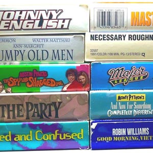 May include: A stack of vintage VHS tapes featuring movie titles like "Johnny English," "Grumpy Old Men," "Austin Powers," and "Dazed and Confused." The tapes have colorful labels and include titles like "Masterminds" and "Good Morning, Vietnam."