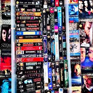 May include: A selection of VHS tapes, mainly horror and thriller films, in a clear plastic container and spread on a light-coloured surface. Titles include "Mimic," "The Cell," and "Urban Legend." The tapes have colourful cover art.