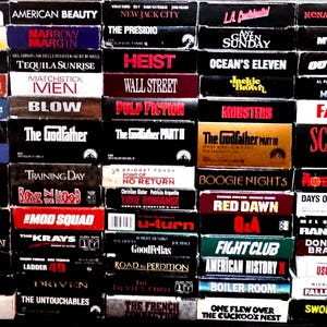 Peut inclure: Une grande collection de boîtiers de films VHS rectangulaires disposés en grille. Les tranches affichent divers titres de films, notamment Snatch, American Beauty, 8 Mile, The Godfather, Pulp Fiction, Scarface, Casino et Fight Club.