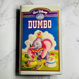 Puede incluir: Una cinta VHS vintage de "Dumbo" de Disney de la colección Masterpiece. La portada presenta una ilustración de dibujos animados de Dumbo el elefante con grandes orejas rosas, un sombrero amarillo y un collar rojo. El título "DUMBO" está en letras rojas grandes.
