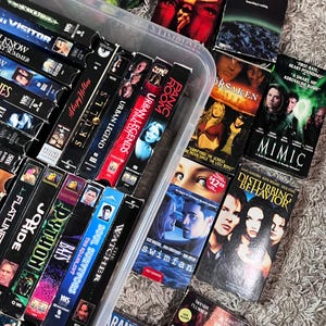Może przedstawiać: Kolekcja r&oacute;żnych kaset VHS przechowywanych w przezroczystym plastikowym pojemniku, zawierająca tytuły takie jak The Dark Side of the Moon, Mimic, Disturbing Behavior, Swimfan, 7 Days to Live, Tarantulas, The Watcher, Soul Survivors, Bats, Python oraz Panic Room.