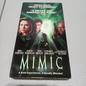 Może przedstawiać: Kaseta VHS z filmem Mimic z zielonkawą okładką przedstawiającą aktor&oacute;w: Mirę Sorvino, Jeremy'ego Northama, Josha Brolina, F. Murraya Abrahama i Charlesa S. Duttona. Tekst głosi First-Rate, Heart-Pounding, A Smart, Hip Adrenaline Rush!